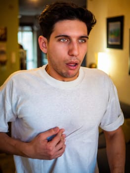 Suffering guy looking at camera and putting hand on chest while feeling heart pain against blurred apartment