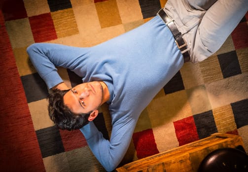 Cool dark-haired handsome young man laying on the floor over colorful rug, looking at camera, hand under his head