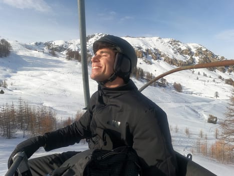 Portrait of handsome man in skiing suit sitting on chairlift rising up and looking at camera.