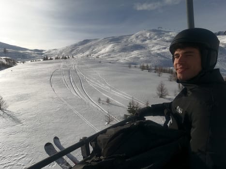 Portrait of handsome man in skiing suit sitting on chairlift rising up and looking at camera.