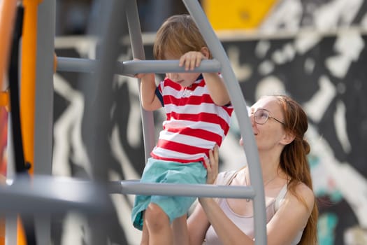 Cute blond boy climbing ladder on playground with support of his smiling mother. Mid shot