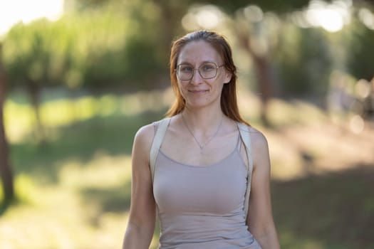 Charming adult woman in glasses with dyed red hair standing in the park. Portrait