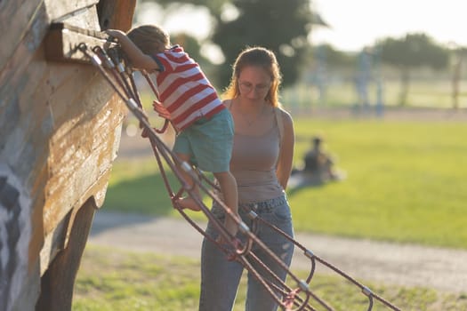 Cute blonde boy climbing the rope ladder on playground and his mother is watching him. Mid shot