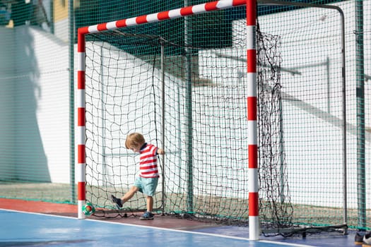 Little blonde smiling boy playing soccer - standing in the gates and hitting back the ball. Mid shot