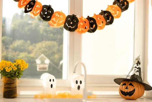 Halloween holiday concept. A pumpkin with a painted face, a white ghost and a bouquet of yellow chrysanthemum flowers in a black vase against the background of a window in home interior.