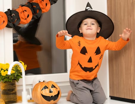 Halloween concept. A small, cheerful, handsome boy in a wizard's hat and an orange sweater sits on a table in the kitchen against the background of a pumpkin, a ghost and a bouquet of yellow flowers. Festive interior.