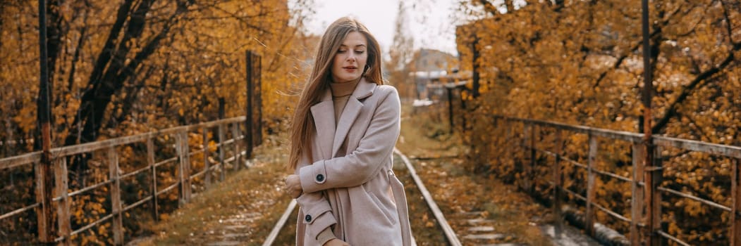 A beautiful long-haired woman walks through the autumn streets. Railway, autumn leaves, a woman in a light coat.