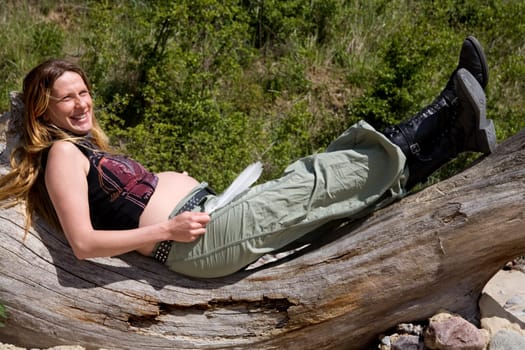 A young pregnant woman lies relaxed on a tree trunk and smiles