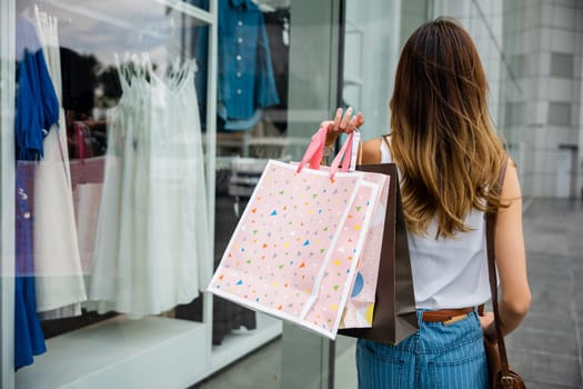 Young woman walking in front of clothing store with shopping bags, surrounded by busy urban buildings and bustling transportation. She is a trendy shopper, always following the latest fashion trends.