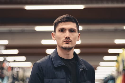 Portrait of one young handsome caucasian guy in a blue jacket looking at the camera while standing in a store, close-up side view.