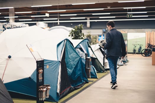One young Caucasian man in a blue jacket stands sideways and examines the tents in the store, close-up side view.
