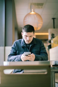 One young Caucasian man in a dark jacket sits at a table by the window in a diner and carefully reads a message in a mobile phone in his hands, close-up view from below.