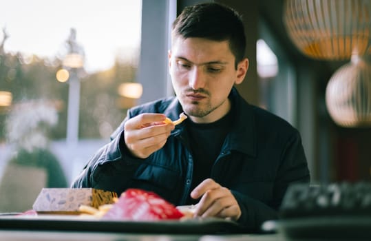 Portrait of one young handsome caucasian brunette man in a dark jacket sits at a table in a diner and appetizingly eats french fries, close-up side view.
