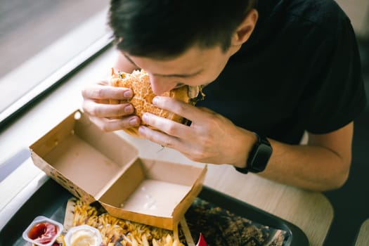 Portrait of one young beautiful caucasian brunette man in a black t-shirt sits at a table near the window in a diner and appetizingly eats a burger, biting it, close-up top view.