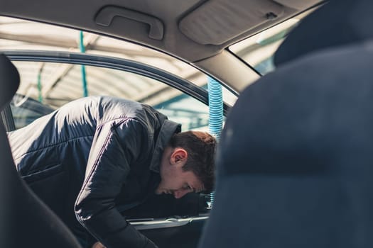 One young handsome caucasian brunette man vacuuming the front seat inside the salon in his car at the city car wash on a clear spring sunny day, side view, close-up.