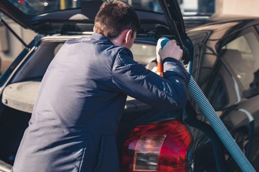 One young handsome caucasian brunette man vacuuming the trunk inside the salon in his car at the city car wash on a clear spring sunny day, top view, close-up.