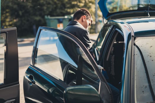 One young handsome Caucasian man in a dark jacket stands sideways near an open car door at a city car wash on a clear spring sunny day, top view, close-up.