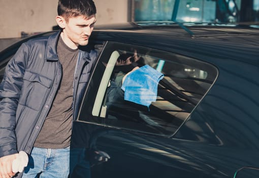 One young handsome Caucasian man in a dark jacket wipes with a blue rag inside the side window of a car at a city car wash on a clear spring sunny day, top view, close-up.