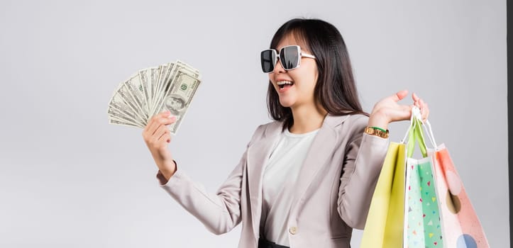 Woman with glasses confident shopper smile holding online shopping bags multicolor and dollar money banknotes on hand, excited happy Asian young female person studio shot isolated on white background