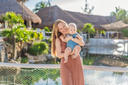 Amidst tropical palms and thatched roofs, a loving mom embraces her baby, sharing warmth and affection in a tranquil outdoor setting.