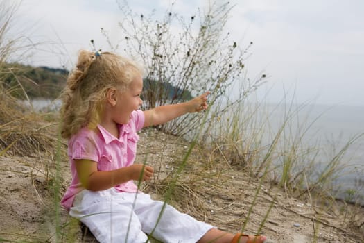 A little girl sits on the beach and points her finger at something