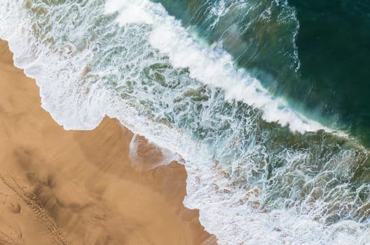 A high-angle shot captures the ocean's powerful waves crashing onto the sandy shore of Lumahai Beach in Kauai, Hawaii.