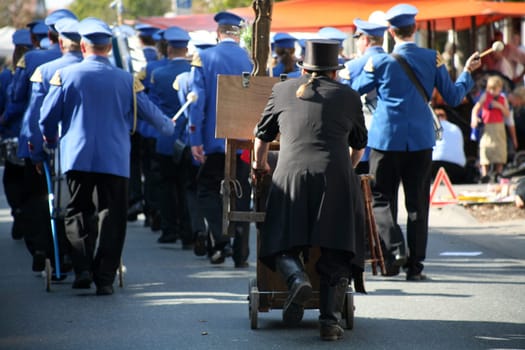 A hurdy-gurdy man dressed in black follows a marching band