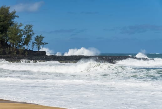Ocean waves crashing on the shore rocks under a blue sky, with the turquoise water rippling and splashing at Lumahai Beach.
