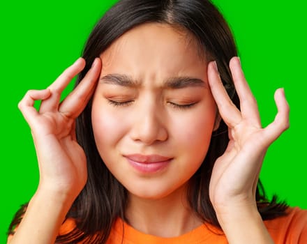 A girl with long dark hair is holding her temples with both hands, showing signs of discomfort. She wears an orange shirt and is set against a vibrant green background.