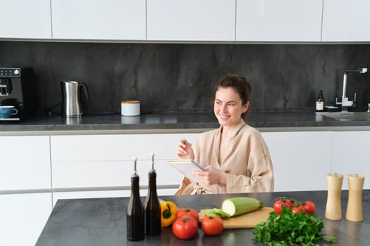 Portrait of woman writing down list of groceries, making notes in recipe, sitting in kitchen near vegetables, preparing dinner menu.