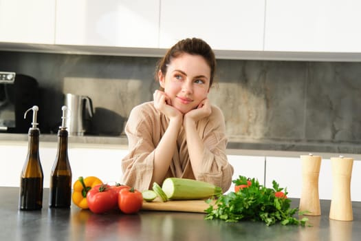 Portrait of smiling young woman leaning on kitchen counter, preparing dinner, standing near chopping board, cutting zucchini, looking away and thinking, thinks what to cook.