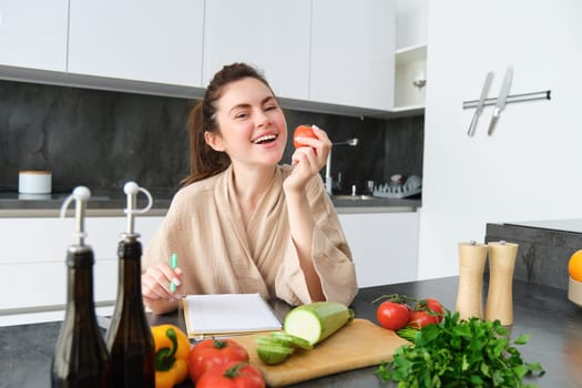 Portrait of happy brunette woman, bites tomato, sits in the kitchen with vegetables, cooking vegetarian meal, preparing list of groceries, dinner menu, wearing bathrobe.