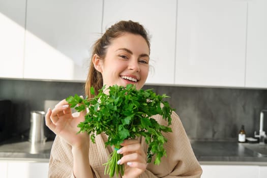 Portrait of beautiful smiling girl with bouquet of parsley, standing in the kitchen and cooking, adding herbs to healthy fresh salad or meal, preparing food.