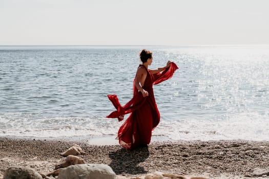 Side view a Young beautiful sensual woman in a red long dress posing on a rock high above the sea during sunrise. Girl on the nature on blue sky background. Fashion photo.