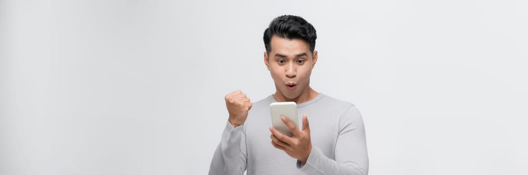 A happy young man holding mobile phone and celebrating success isolated over white background