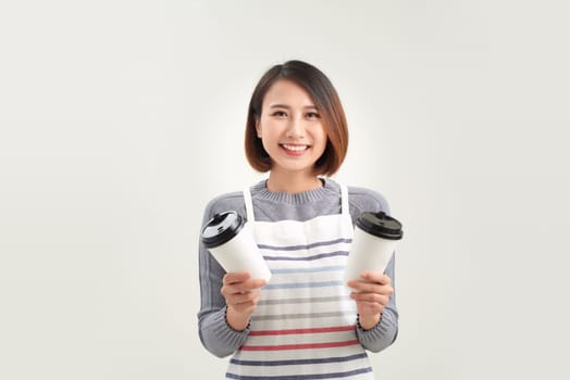 Friendly asian young woman in apron holding takeaway paper coffee cup