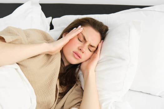 Woman lying in bed and feeling unwell, having headache, touching her head with frustrated face, has high fever or migraine. concept of health and people.