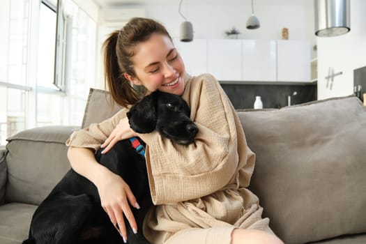 Animals and lifestyle concept. Happy young woman in bathrobe, hugs her dog on sofa, cuddle puppy and smiling.