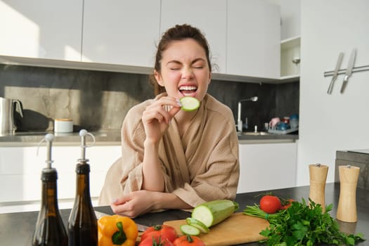 Portrait of happy girl eating vegetables while making meal, cooking healthy vegetarian food in kitchen, holding zucchini, posing in bathrobe.