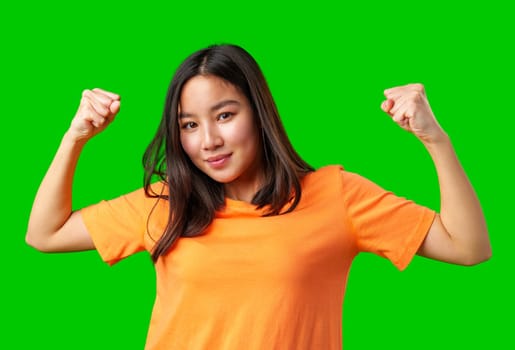 A young girl expresses confidence by flexing her arms while wearing an orange shirt. The bright green background emphasizes her positive energy and spirit.