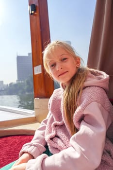 A girl relaxes on a boat, dressed in a cozy pink outfit, looking at the camera. The sunny city skyline and calm waters create a serene atmosphere around her.