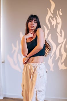 A model strikes a confident pose in a black crop top and light cargo pants. The studio features artistic flame shadows reflecting on the wall, enhancing the creative atmosphere.