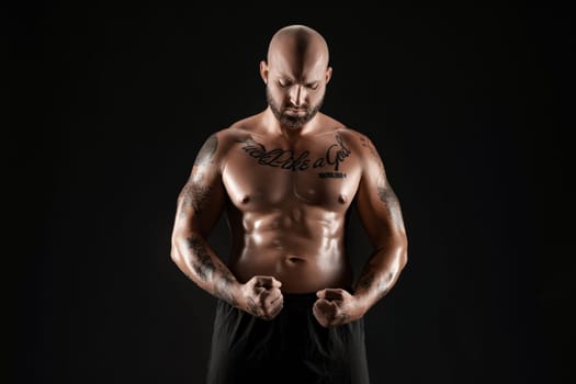 Good-looking bald, bearded, tattooed man in black shorts is demonstating his muscles posing against a black background and looking down. Chic muscular body, fitness, gym, healthy lifestyle concept. Close-up portrait.