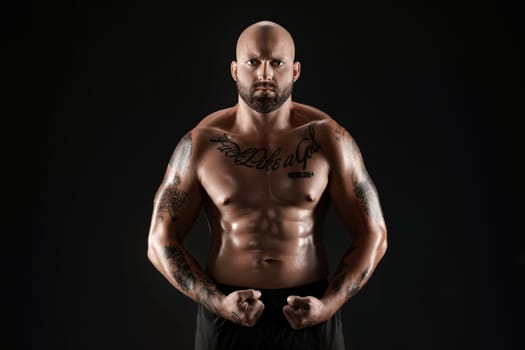 Good-looking bald, bearded, tattooed male in black shorts is demonstating his muscles posing against a black background and looking at the camera. Chic muscular body, fitness, gym, healthy lifestyle concept. Close-up portrait.