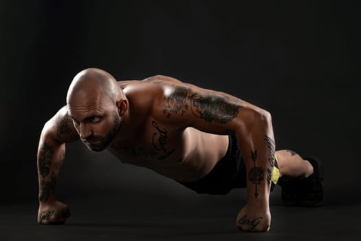Stately bald, bearded, tattooed male in black shorts and sneakers is pushing ups from the floor against a black background. Chic muscular body, fitness, gym, healthy lifestyle concept. Close-up portrait.