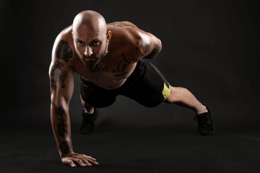 Stately bald, bearded, tattooed guy in black shorts and sneakers is pushing ups from the floor against a black background. Chic muscular body, fitness, gym, healthy lifestyle concept. Close-up portrait.