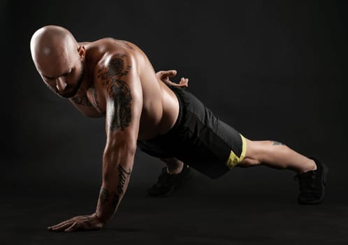 Stately bald, bearded, tattooed person in black shorts and sneakers is pushing ups from the floor against a black background. Chic muscular body, fitness, gym, healthy lifestyle concept. Close-up portrait.