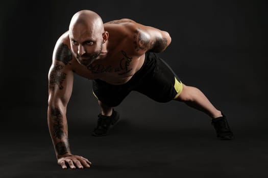 Stately bald, bearded, tattooed fellow in black shorts and sneakers is pushing ups from the floor against a black background. Chic muscular body, fitness, gym, healthy lifestyle concept. Close-up portrait.