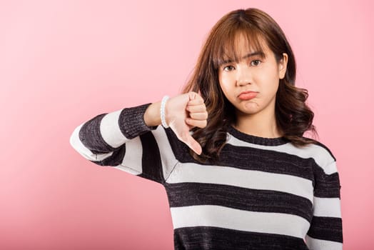 An Asian woman, captured in a portrait, reveals her displeasure with a thumbs-down gesture, indicating rejection. Studio shot isolated on pink, highlighting her negative expression.