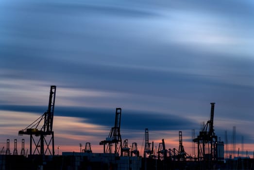 "Long-exposure photo captures a busy port at dusk, with cranes silhouetted against a colorful sky, symbolizing global trade's relentless pace."